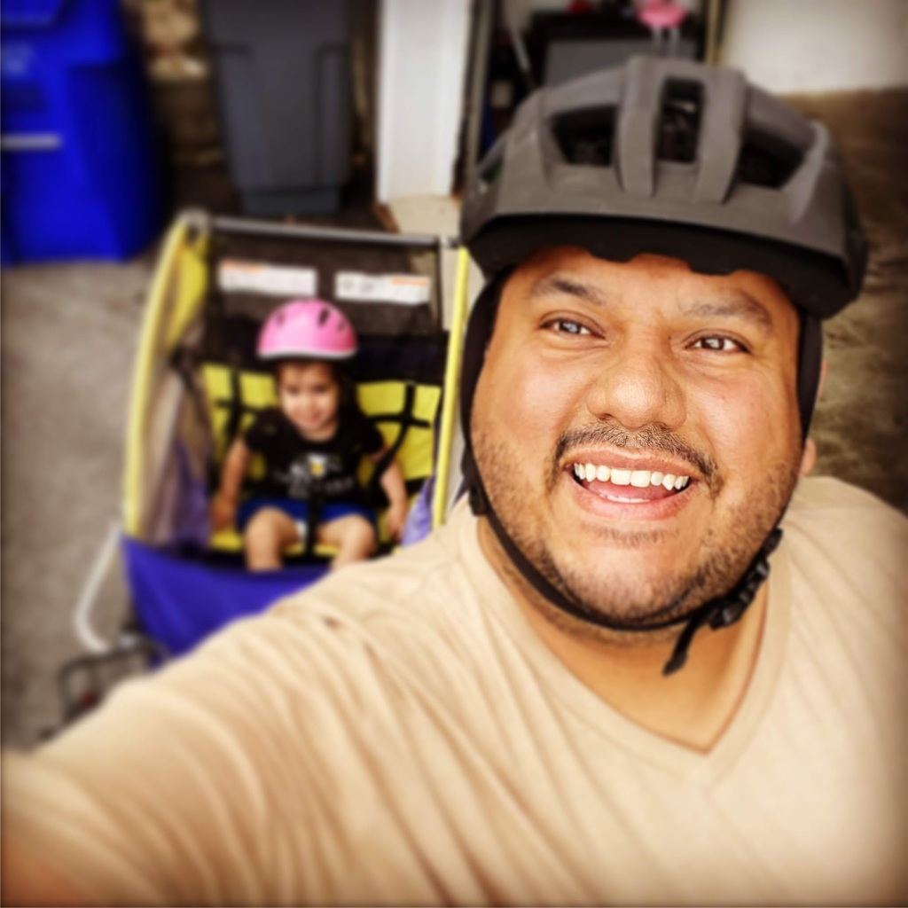 Image of a man wearing a bicycle helmet and smiling at the camera. Behind him is a little girl with her own pink bicycle helmet and smiling at the camera as well.