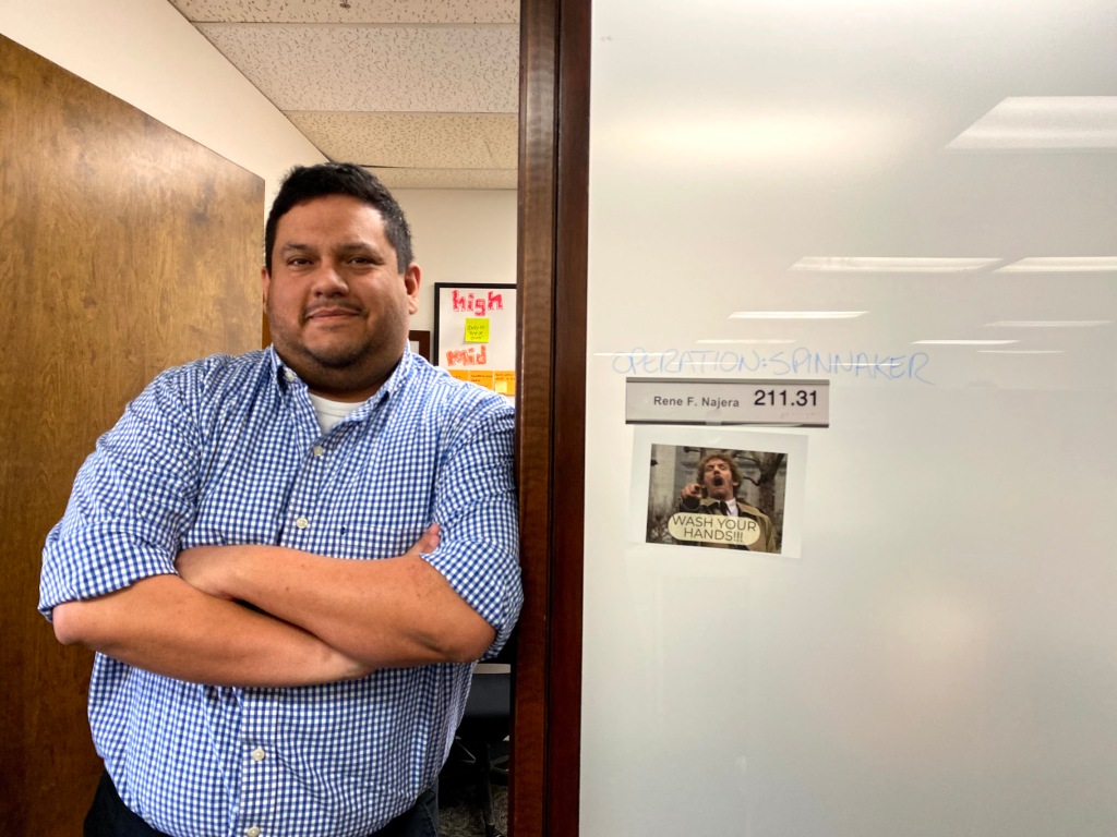 Image of a man standing at the threshold to a door. He has his arms crossed and is smiling at the camera. He is wearing a blue shirt. The sign to his door reads "Operation Spinnaker" and "Rene F Najera, 211.31" and there is an image taped to the side of the door with a man pointing at the camera and a caption reading, "Wash your hands!"