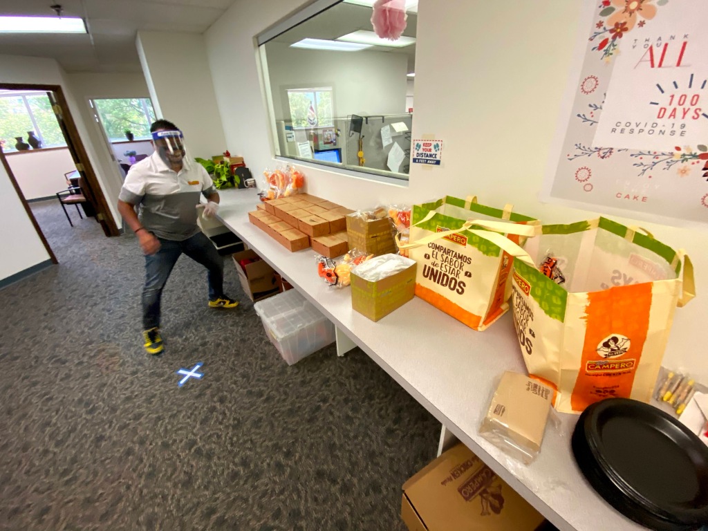 Image of a man standing in front of several boxes of food sitting on a table. He is wearing a face shield and a mask.