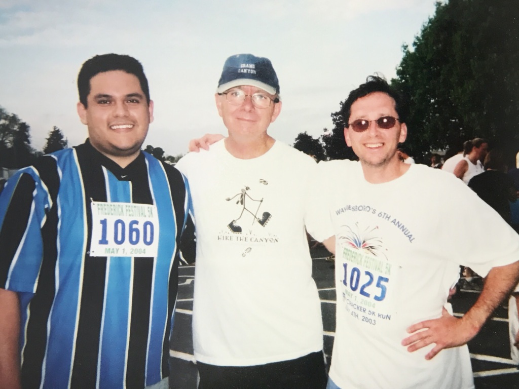 Image of three men standing together and smiling at the camera as they're wearing racing bibs and sports clothing.