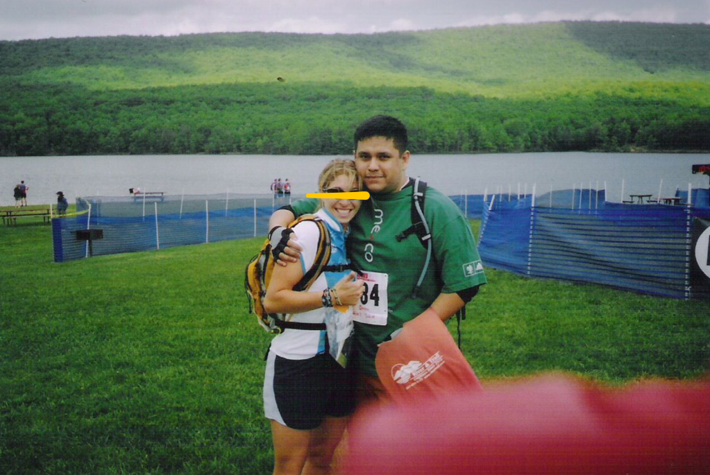 Image of a man and a woman in an outdoor setting. He is hugging her as she shows a thumbs up sign. He is wearing a green shirt and bicycle gloves while she is wearing a white shirt. They both have backpacks on, and a lake can be seen in the background.