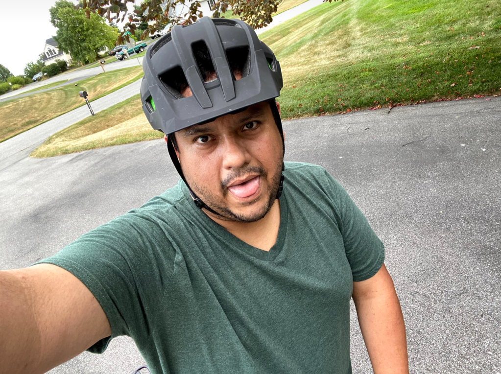 Image of a man wearing a bicycle helmet and a green shirt. He is looking at the camera and sticking out his tongue.