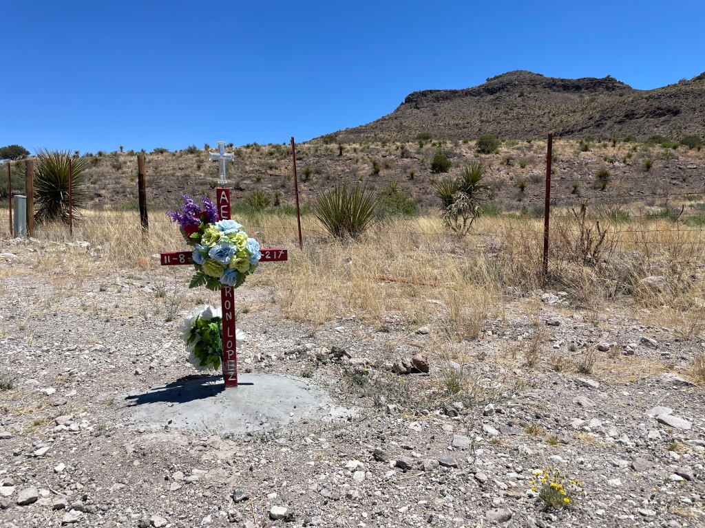 A memorial in the shape of a cross with floral arrangement on the side of the road with a desert background.