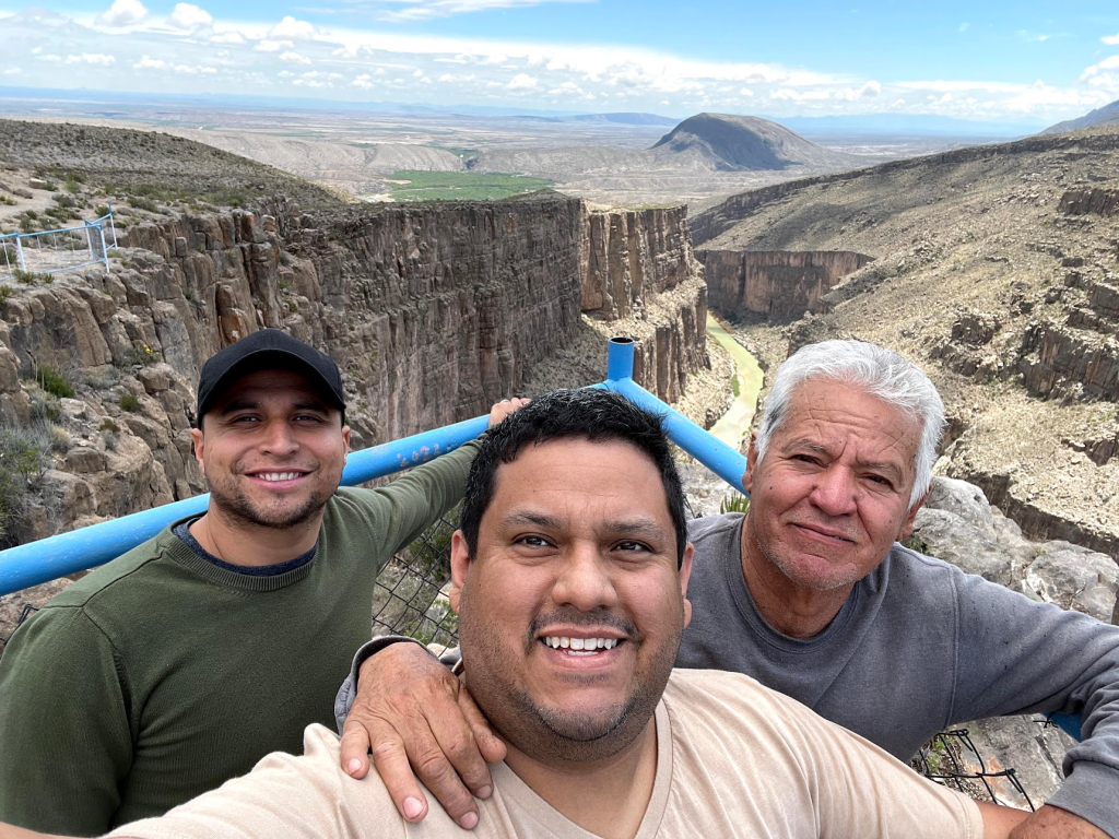 Three men stand in the foreground as a canyon can be seen behind them. The oldest man on the right has his hand on the shoulder of the man in the middle. All three are smiling.