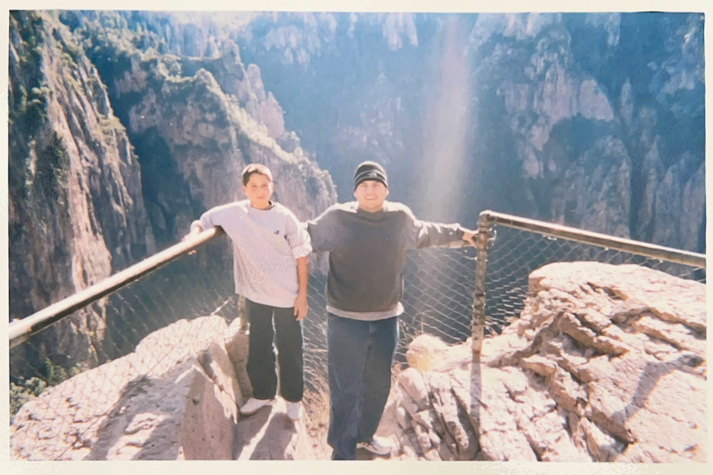 Two young men stand in front of a fence while a chasm and mountains rise behind them.