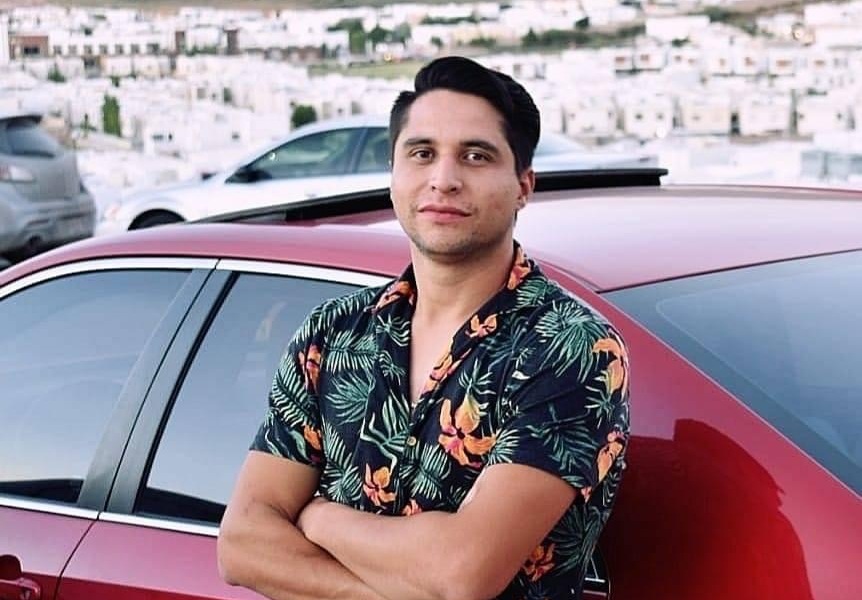 A young man leans back on a red car as he looks at the camera