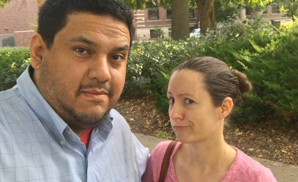 A couple looks at the camera with strange expressions on their face. There is green shrubbery in the background.