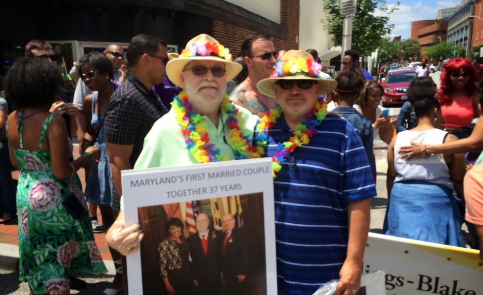 Two men stand in front of a crowd, looking at the camera. One of them holds a large picture with a caption that reads "Maryland's First Married Couple. Together 37 years."