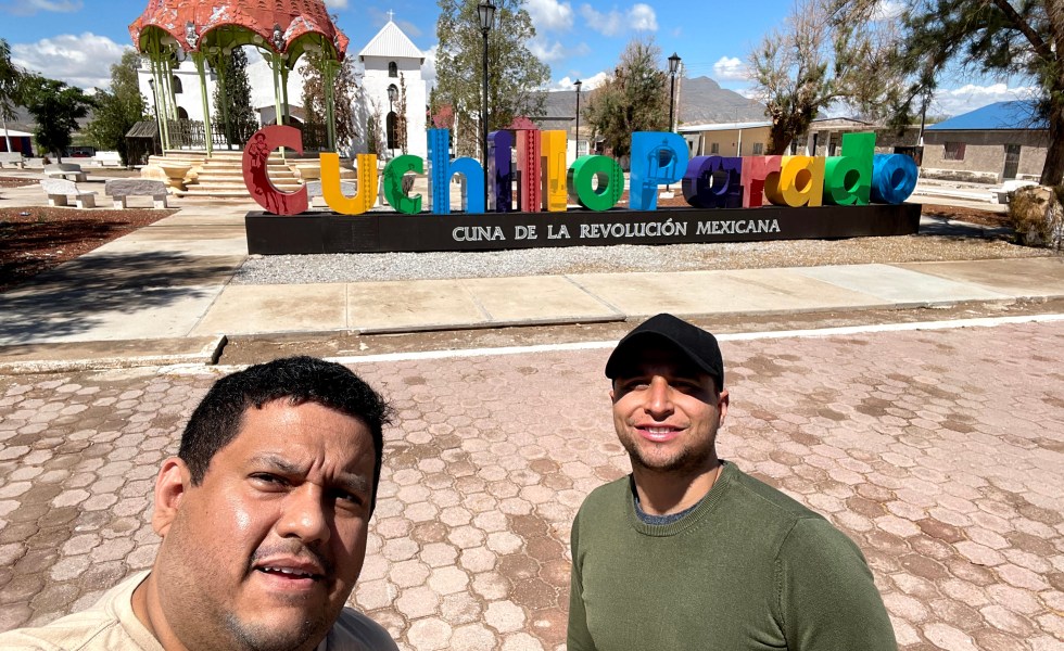 Two men in front of a sign in a small town in northern Mexico. One wears a green shirt and hat. The other wears a tan shirt.
