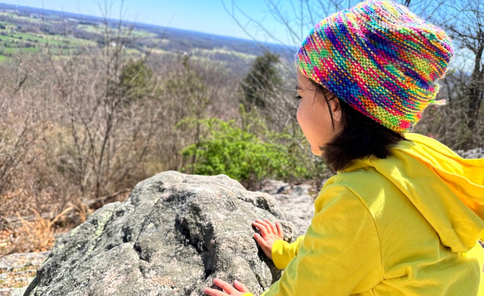 A little girl with a yellow hoodie looks over a valley from a rocky formation on a mountain.