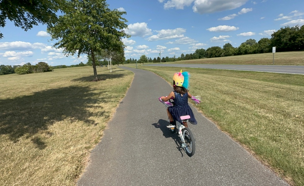 A little girl in a blue dress rides a pink bicycle on an asphalt trail at a park.