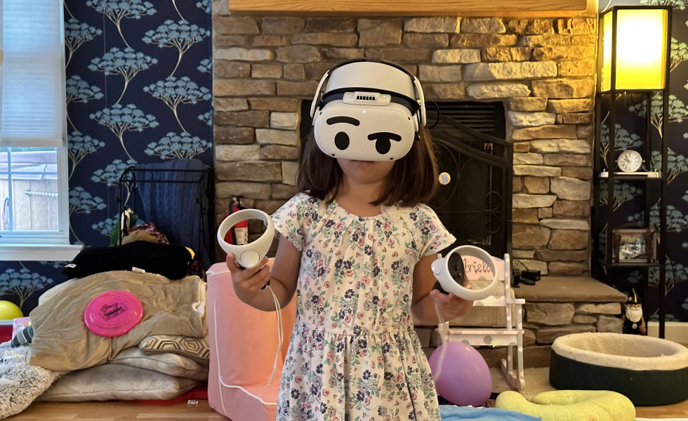 A little girl plays with a virtual reality headset while holding two controllers. Behind her are toys and cushions on the floor.