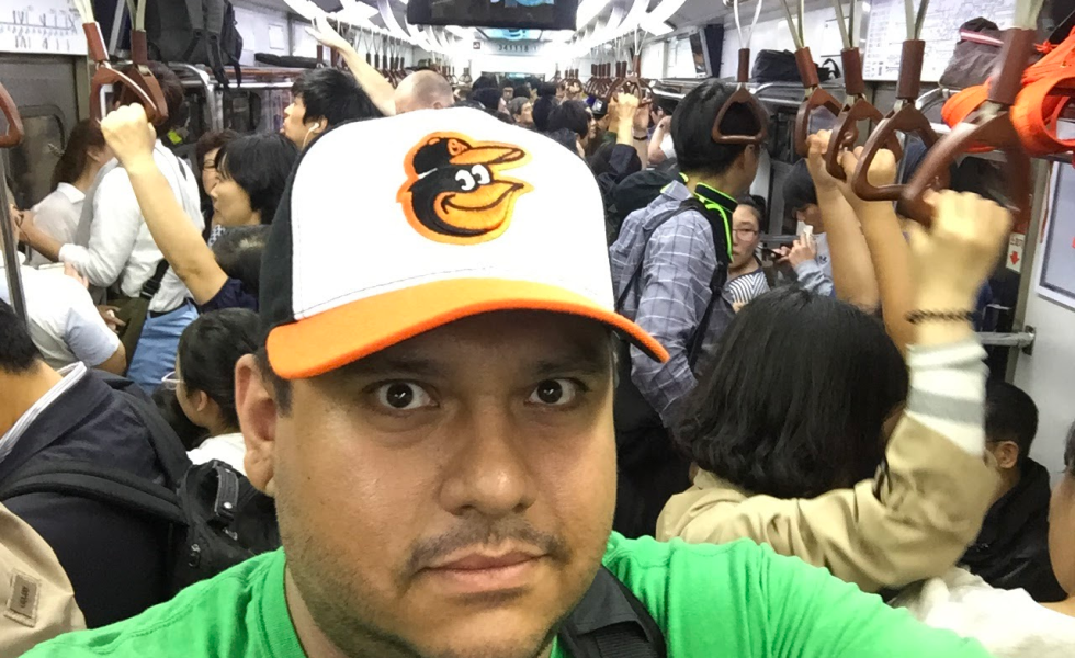 A man with a Baltimore Orioles baseball hat looks at the camera while riding a metro train filled with passengers.